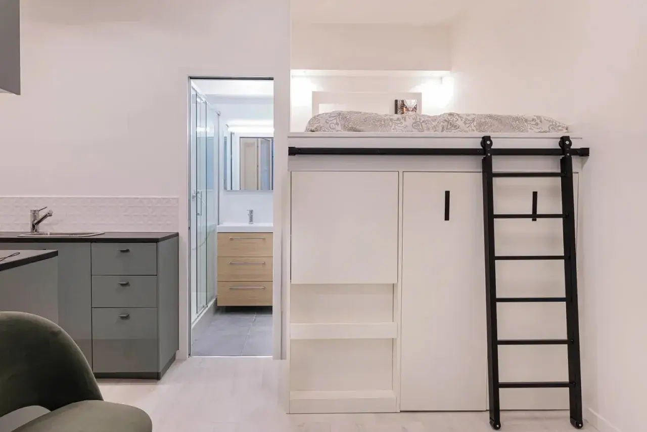 Un appartement fonctionnel avec lit mezzanine, échelle noire, cuisine grise et salle de bain visible.